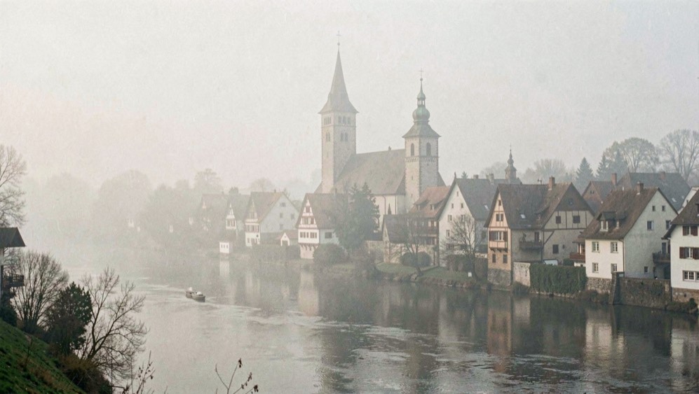 Kirchturm und Satteldächer des Dorfes von der anderen Flussseite im Nebel des frühen Morgens