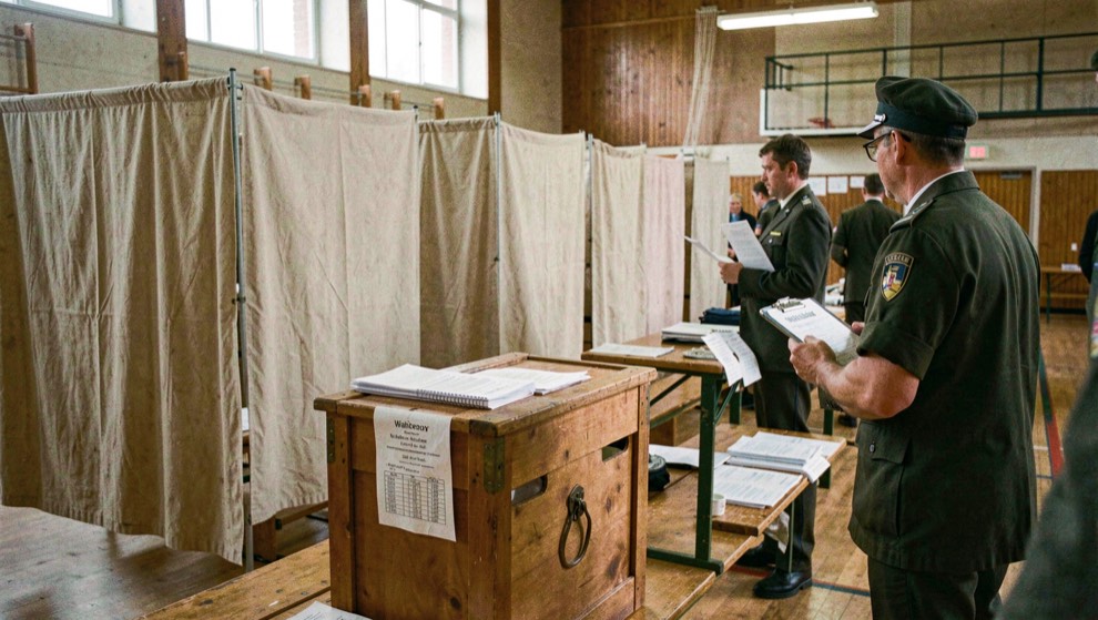Wahllokal in der Turnhalle: Stimmkabinen aus Stoff, Wahlurne und Helfer mit Wählerverzeichnis